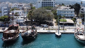 A marina with several boats docked, a modern building labeled "PASMINE HOTEL" in the background, and a cityscape with buildings and greenery. - Jasmine Hotel Apartments (Kos)