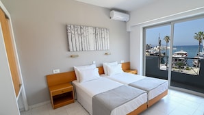 A hotel room with two beds, a large window with a view of the sea and boats, a wall-mounted air conditioner, and a wooden nightstand. - Jasmine Hotel Apartments (Kos)