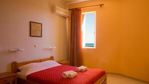 A single bed with a red quilt, two rolled towels, a small bedside table, a wall-mounted air conditioner, a window with curtains, and a framed picture on the wall. - Sunrise Hotel (Chios)