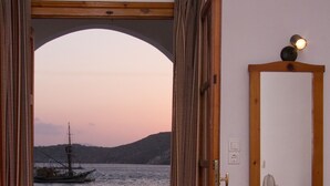 Una camera con un letto, una cassettiera in legno, uno specchio e un tavolo con sedie. Si vede il mare e una barca fuori dalla porta.