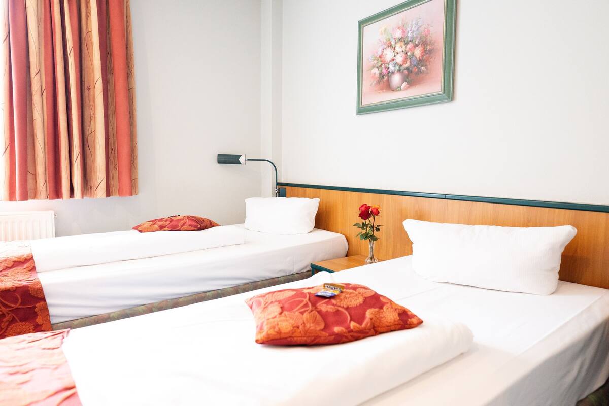 A hotel room with two single beds, a wooden headboard, a bedside table with a vase of flowers, and a framed floral artwork on the wall.