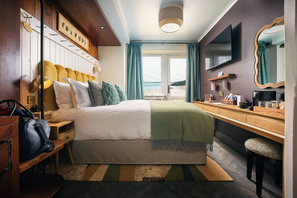 A hotel room with a bed, bedside table, dresser, mirror, and a view of the beach.