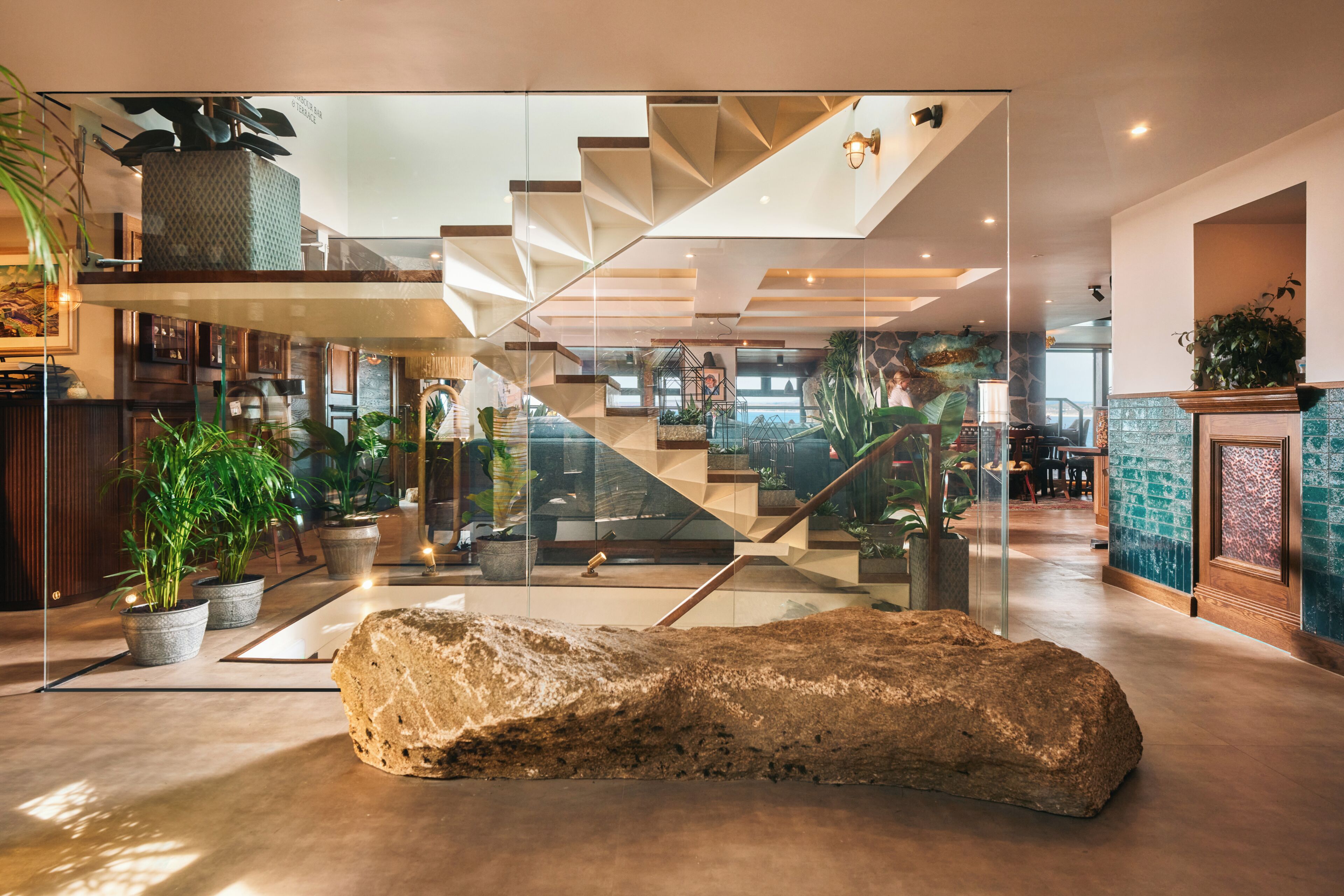 A modern interior with a central glass staircase, potted plants, and a large rock centerpiece.