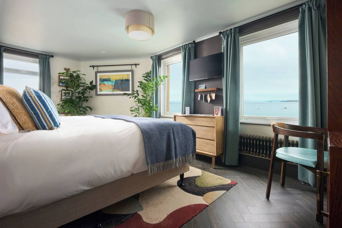 A bedroom with a bed, a dresser, a chair, a TV, and a view of the sea.