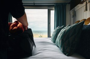 Una cama con vista a la playa, una ventana con cortinas y una persona sosteniendo un bolso.
