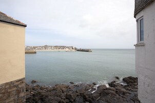 Vista costera con un litoral rocoso, un muelle que se adentra en aguas tranquilas y un pueblo distante con edificios.