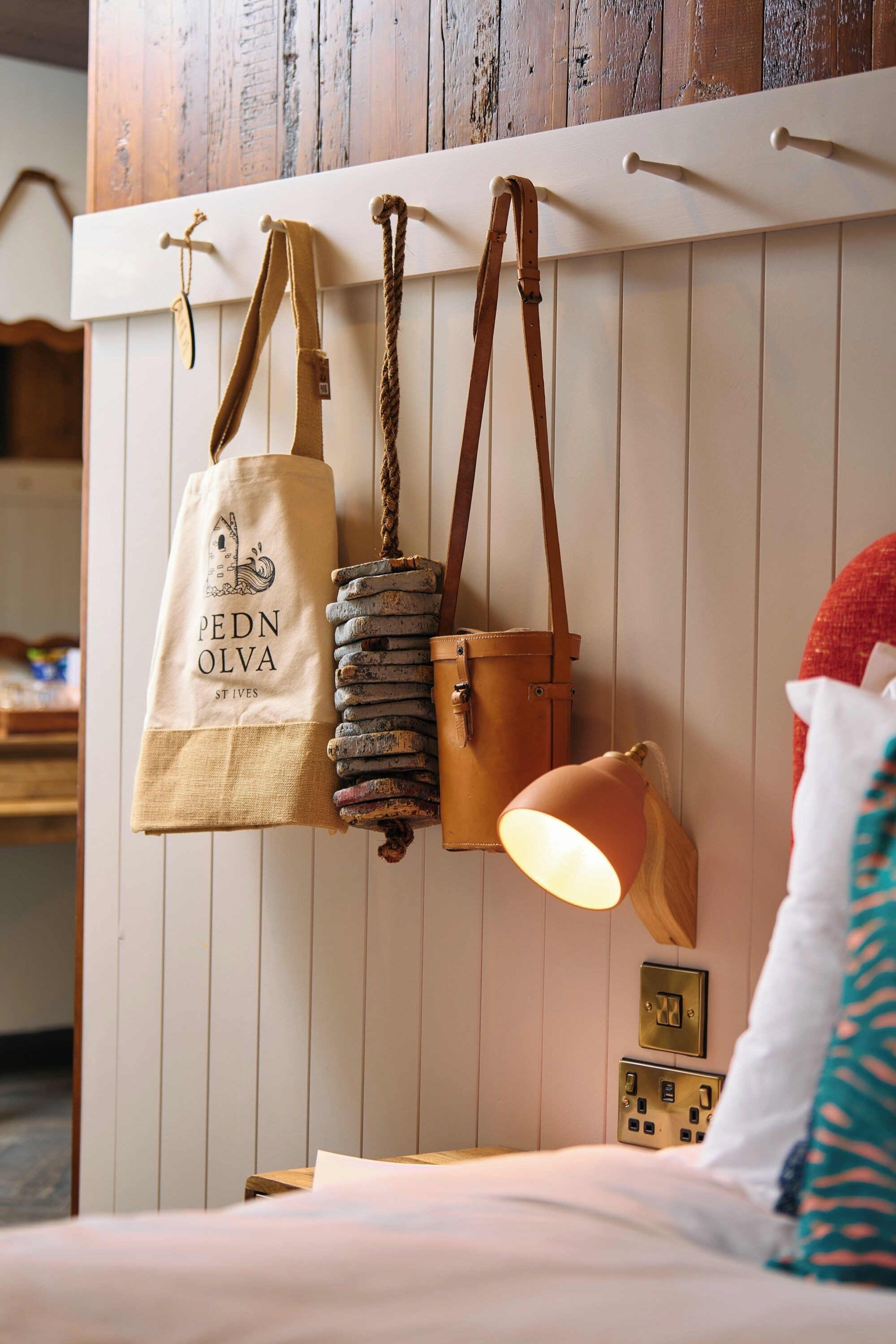 Un lit avec une lampe, une étagère fixée au mur avec des sacs, et une prise de courant.