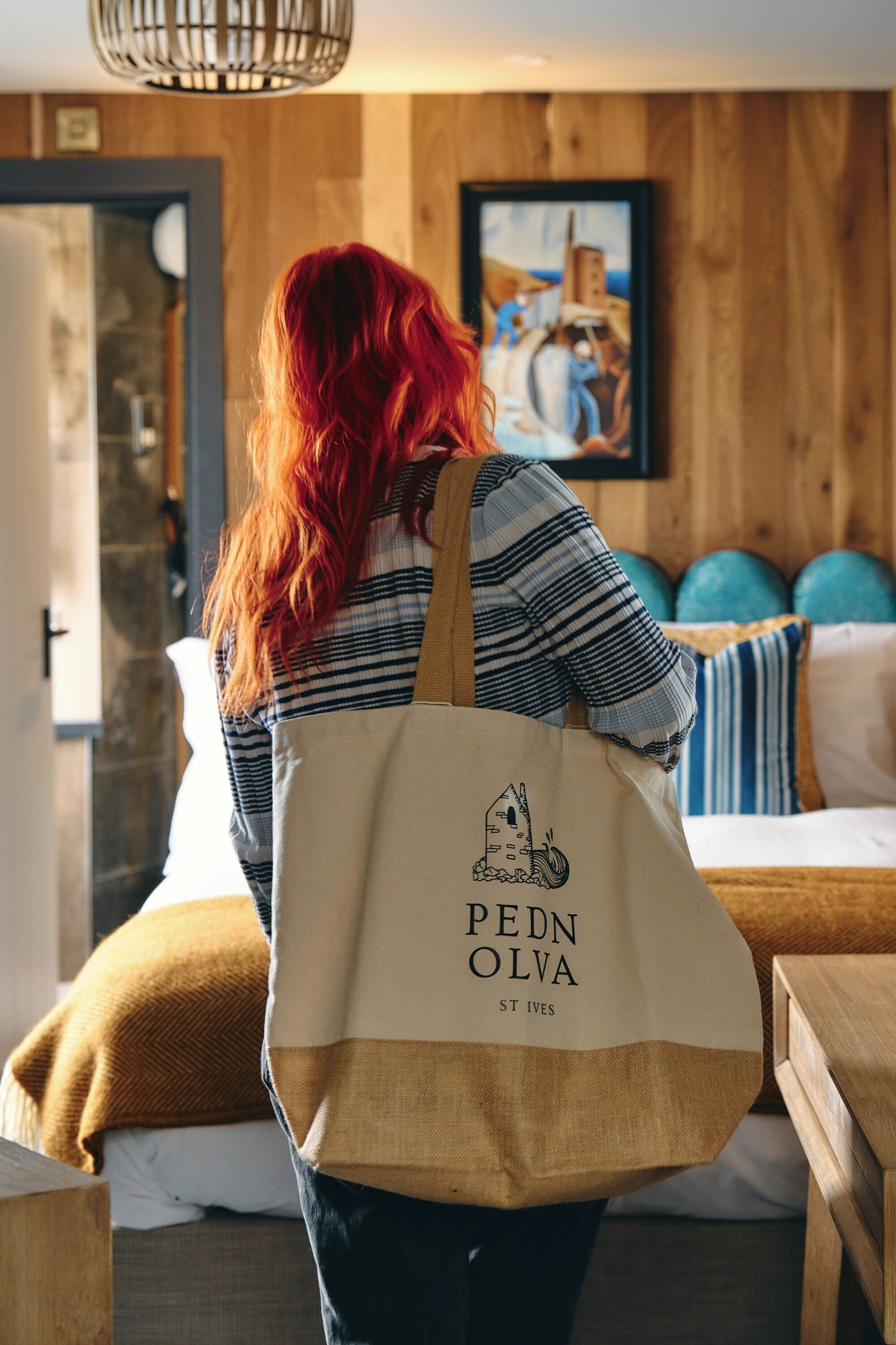 A person with red hair wearing a striped shirt and a beige tote bag with 'PEDN OLVA ST IVES' printed on it.
