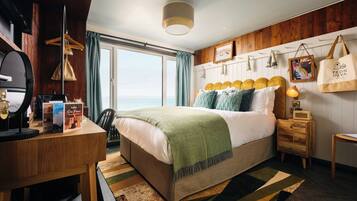 Una habitación de hotel con cama, escritorio, silla, lámpara, marco de fotos y vista al mar.
