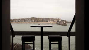 Vista desde una ventana con dos taburetes, que da a un pueblo costero con edificios, un faro y barcos en el agua.