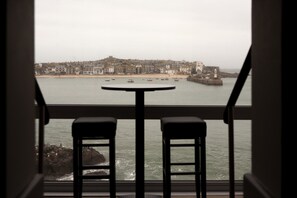 A view from a window with two bar stools, overlooking a coastal town with buildings, a lighthouse, and boats on the water. - Pedn Olva (St Ives)