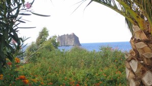A coastal landscape with a rocky outcrop in the background, surrounded by dense vegetation and flowering plants. - Pensione La Nassa (Lipari)