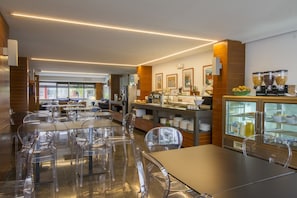 A modern breakfast area with clear chairs, a long table, and a refrigerated display case with various food items.
