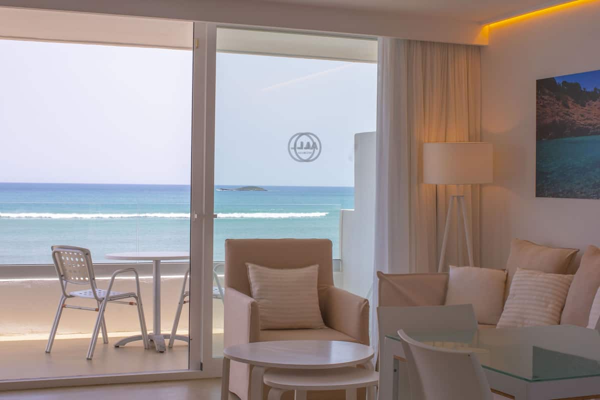 A hotel room with a balcony overlooking the ocean, featuring a sofa, a table, and a chair.