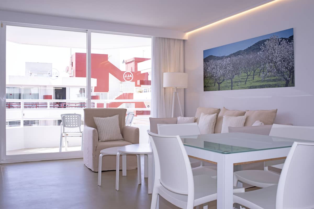 A modern hotel room with a balcony, a dining area, and a large window offering a view of the cityscape.