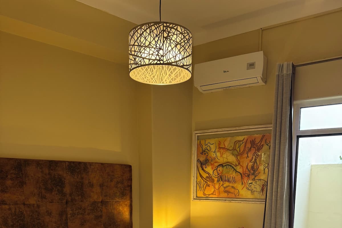 Habitación de hotel con una cama grande, cabecero, lámpara de noche, una mesita con lámpara, un cuadro en la pared y una ventana con vistas a un área verde exterior.