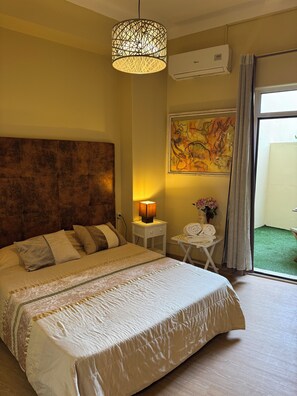 Habitación de hotel con una cama grande, cabecero, lámpara de noche, una mesita con lámpara, un cuadro en la pared y una ventana con vistas a un área verde exterior.