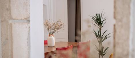 Un coin repas avec une table en bois, des chaises rouges, un vase blanc contenant des fleurs séchées et une plante en pot.