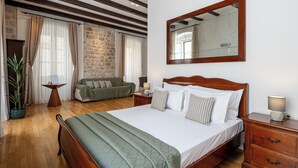 A bedroom with a wooden bed, a nightstand, a bedside table, a mirror, a green armchair, a stone wall, and a potted plant. - Blasius Residence (Dubrovnik)