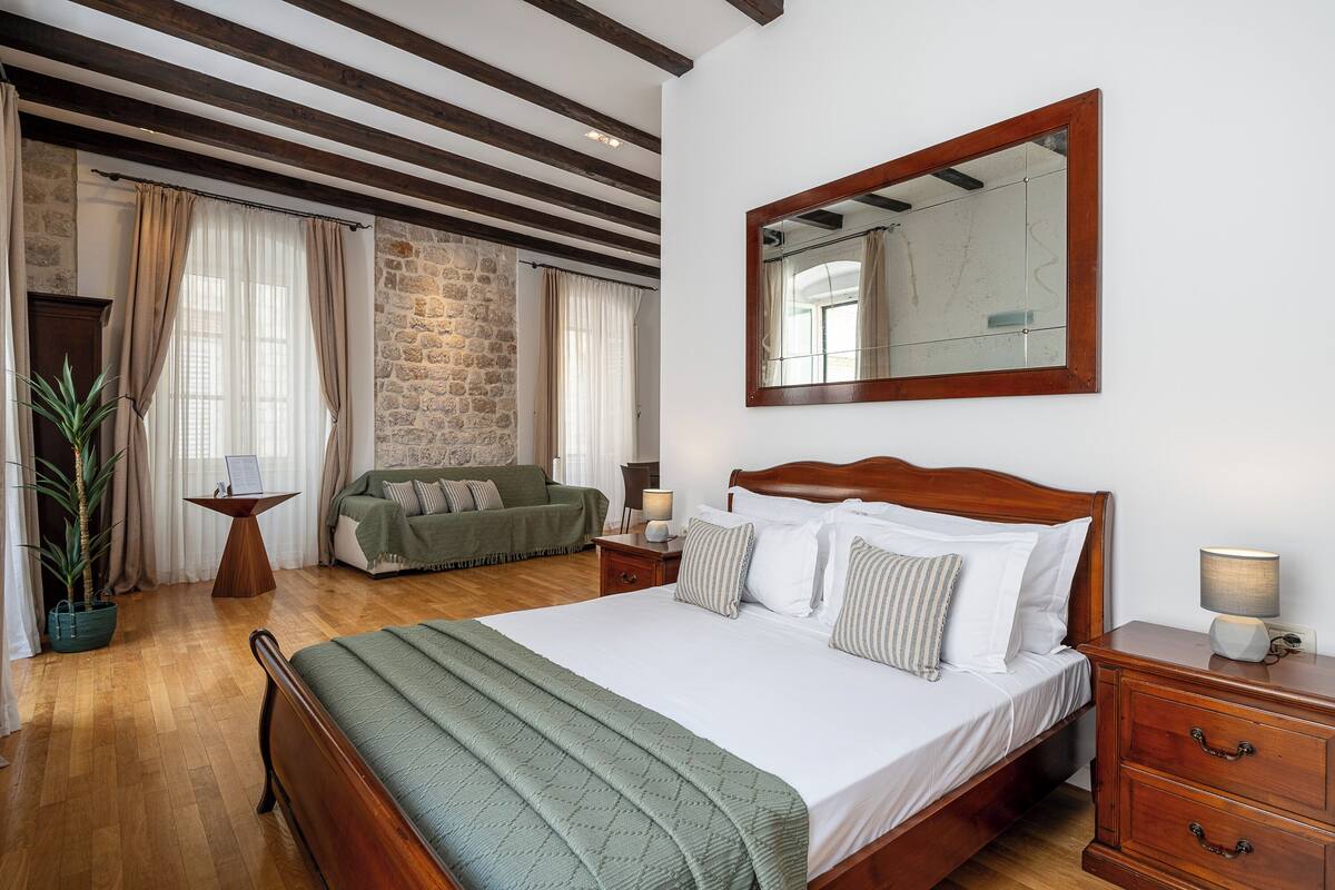 Un dormitorio con cama de madera, mesita de noche, espejo, sillón verde, pared de piedra y planta en maceta.