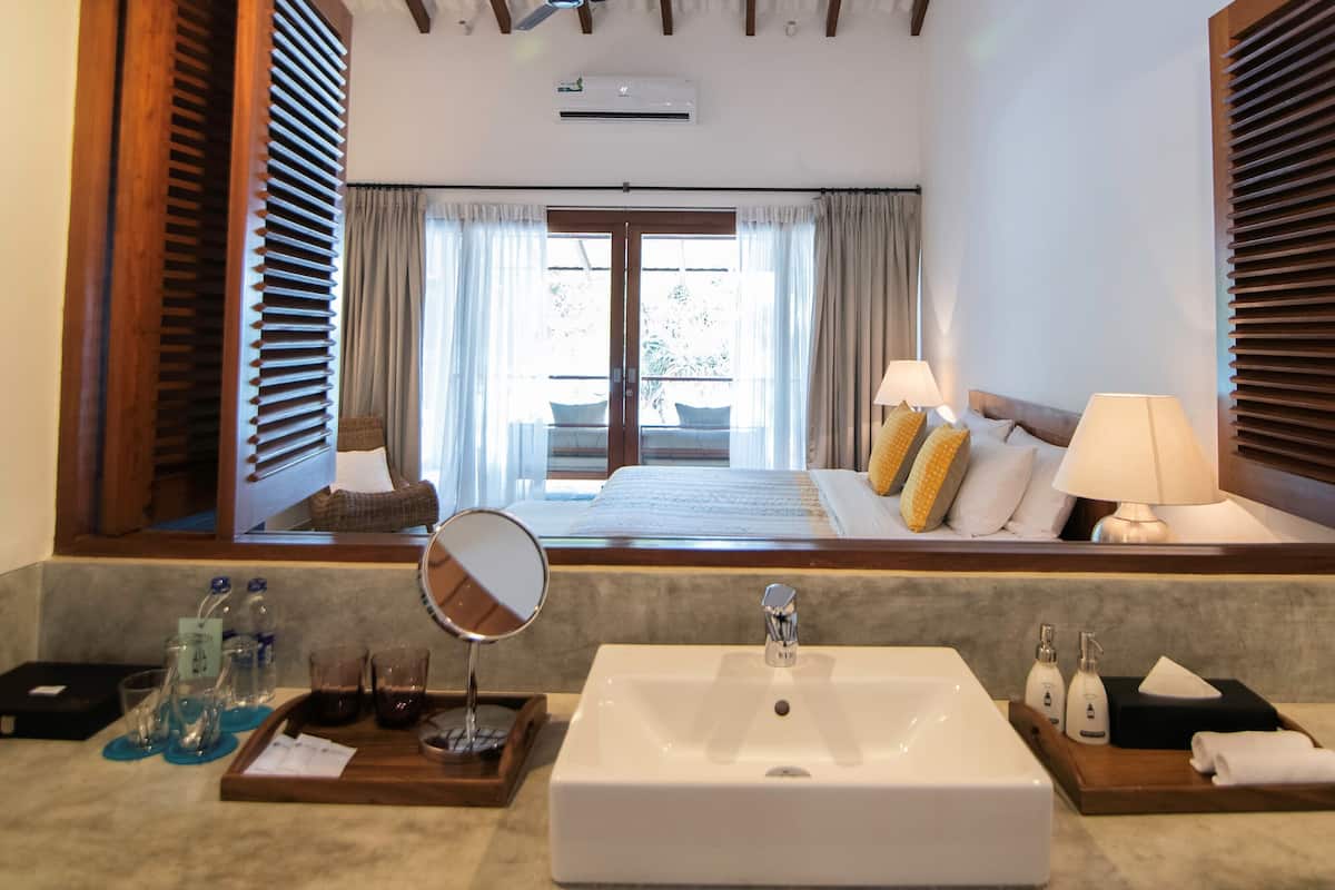 Habitación de hotel con un espejo grande, un lavabo blanco, una mesita de noche con artículos de aseo, una cama con almohadas y vistas a un balcón por la ventana.