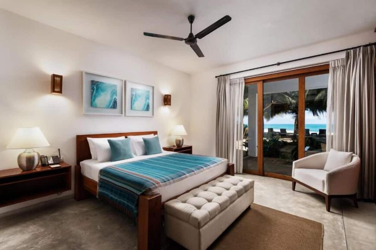 Habitación de hotel con cama, una silla, un ventilador de techo y vistas a la playa a través de una puerta corrediza de vidrio.
