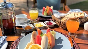 Un desayuno en la playa con una mesa dispuesta para dos personas, que incluye un plato de fruta, un vaso de zumo de naranja y una cafetera.