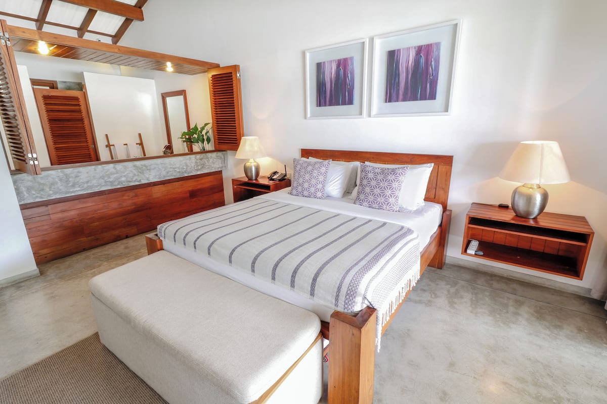 Un dormitorio con una cama grande, cabecera de madera, mesitas de noche con lámparas, un espejo y dos cuadros en la pared.