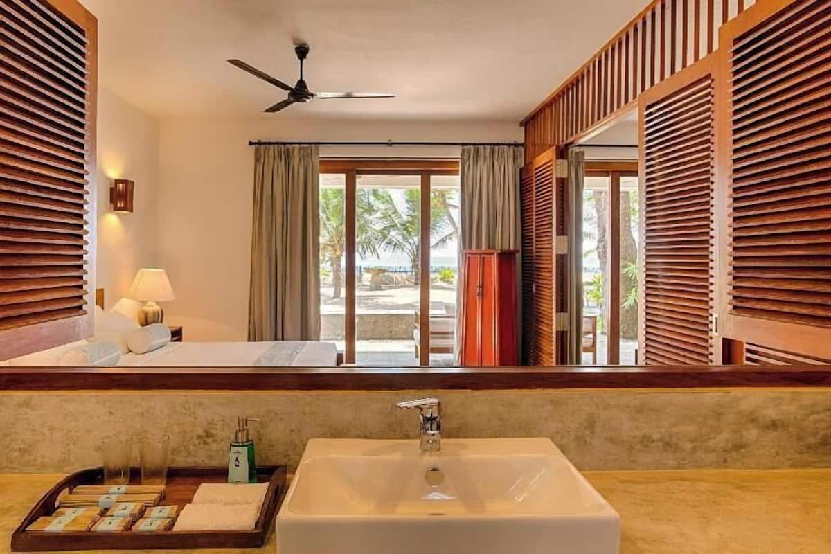 Habitación de hotel con un ventanal grande, ventilador de techo, lavamanos y mesita de noche con lámpara.