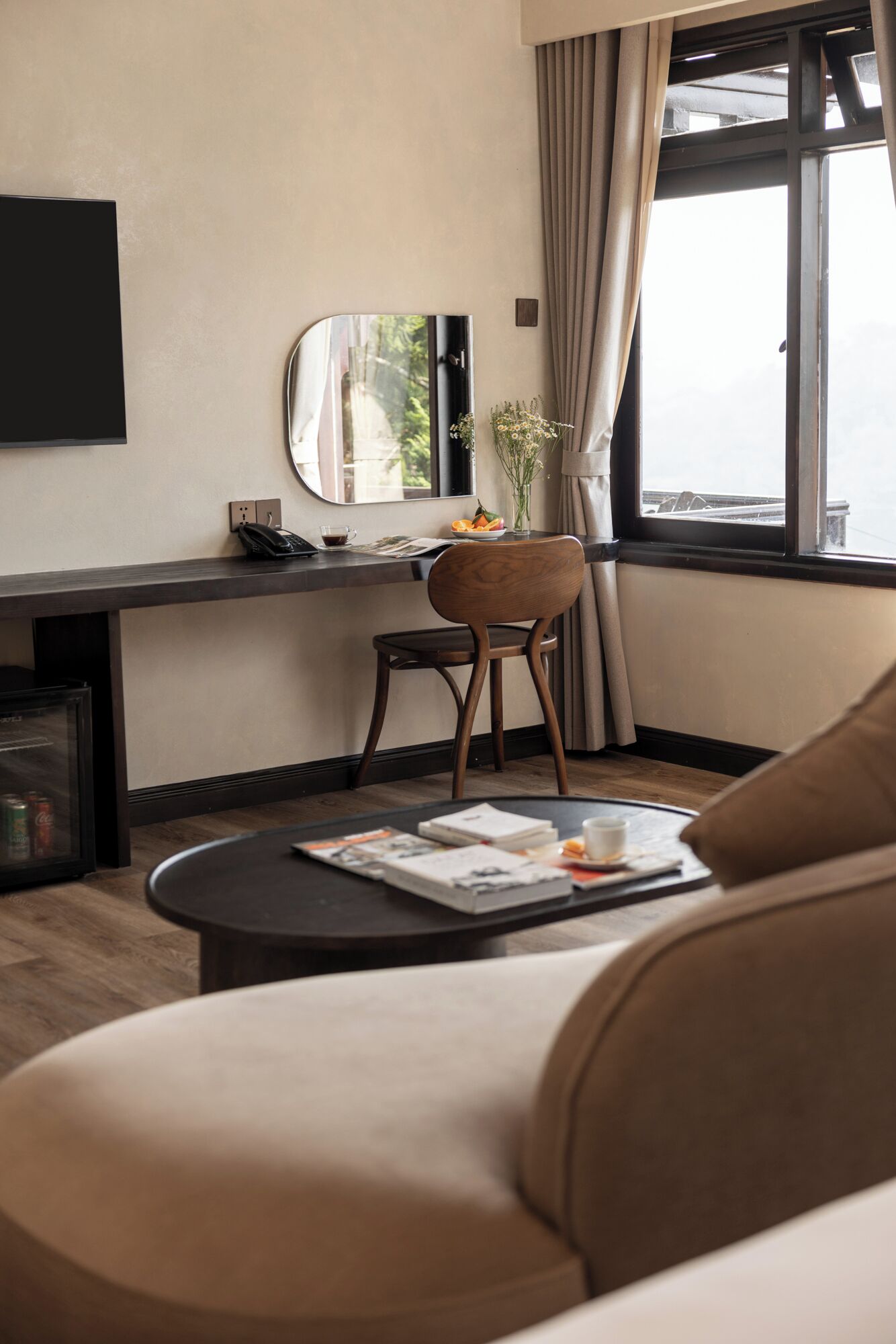 Une chambre d’hôtel moderne avec un canapé, une petite table ronde sur laquelle repose un magazine, une chaise, un bureau avec un miroir et une fenêtre avec des rideaux.