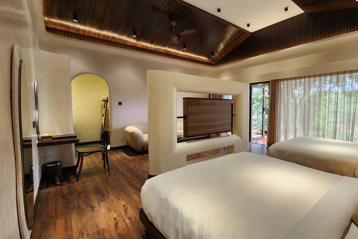 A bedroom with a large bed, a TV mounted on a wall, a desk with a chair, and a window with curtains.