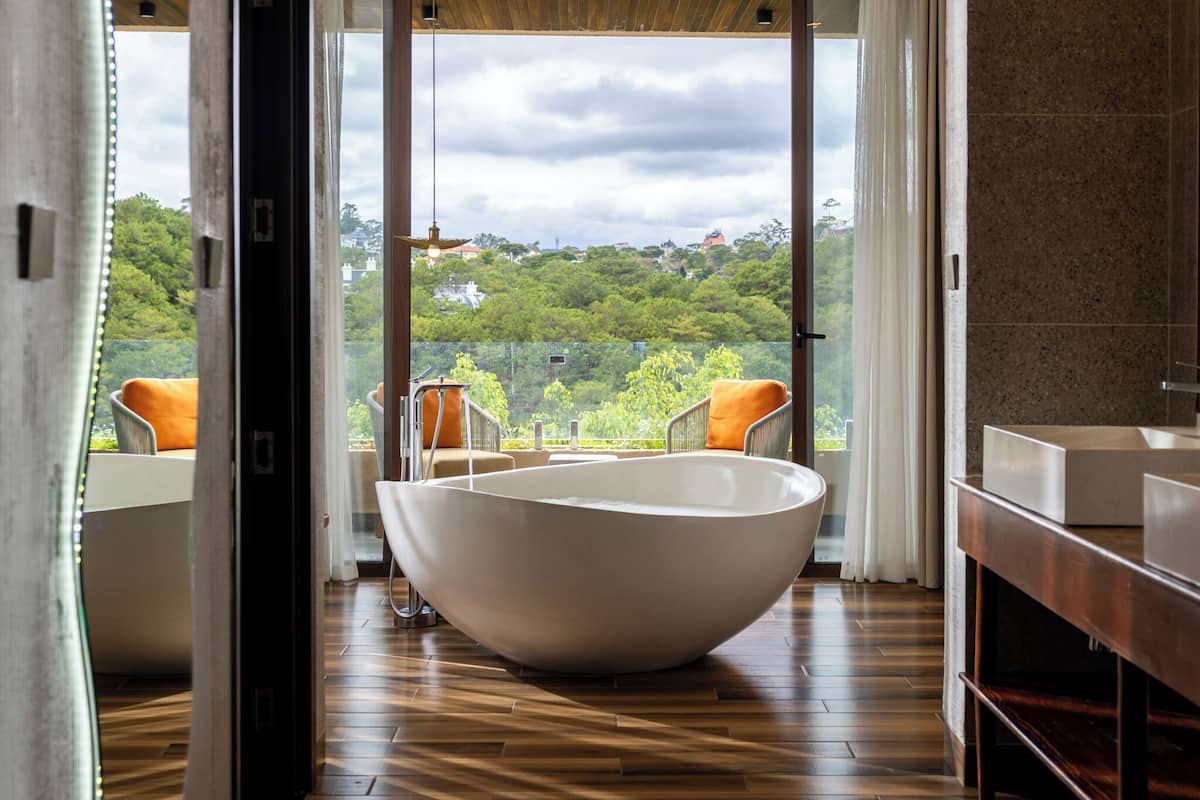 A modern bathroom with a large white bathtub, wooden flooring, and a view of a lush green landscape through large windows.