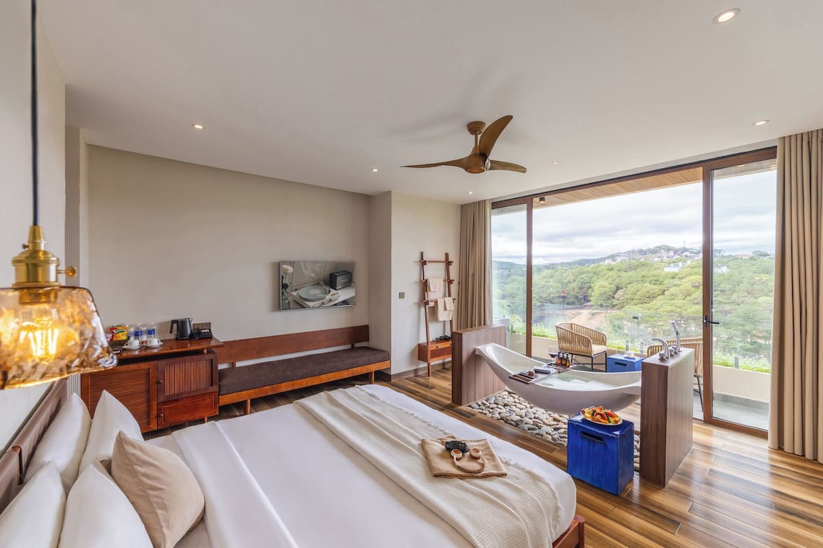 A modern hotel room with a large bed, a sitting area, a bathtub, and a balcony with a city view.