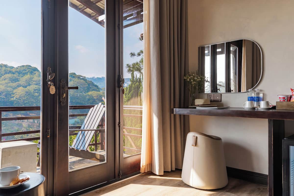 A hotel room with a balcony, a desk with a mirror, a heater, and a view of a forested area.