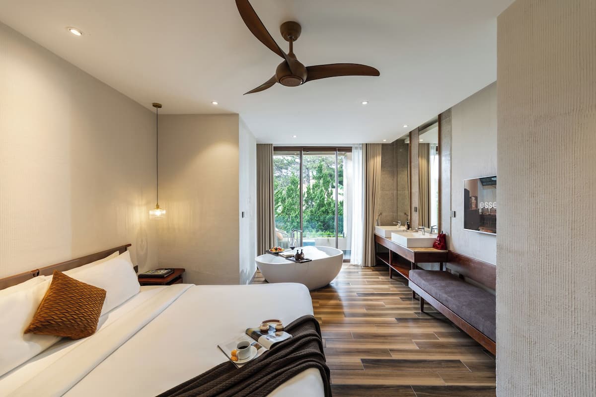 A modern hotel room with a large bed, a ceiling fan, a bathtub, a vanity with a mirror, and a view of greenery outside.