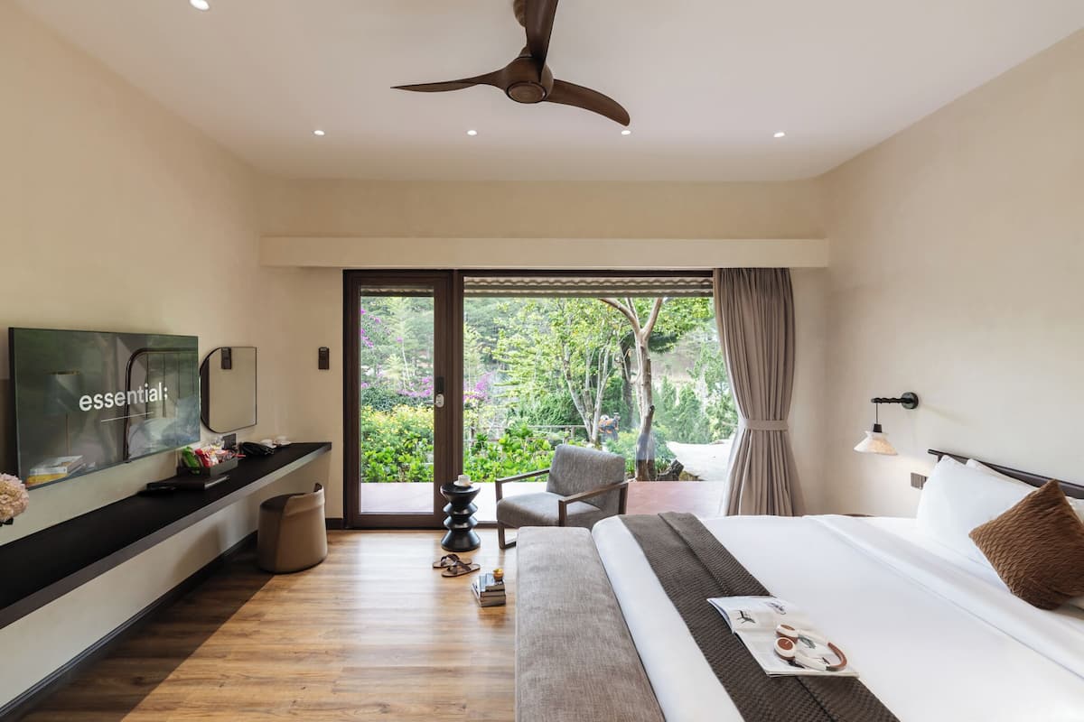 A modern hotel room with a bed, a TV, a desk with a computer, a chair, and a view of greenery through large windows.