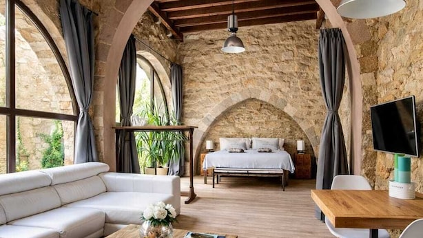 Un interior rústico con pared de piedra, vigas de madera, un sofá, una cama, una mesa de comedor y un televisor.