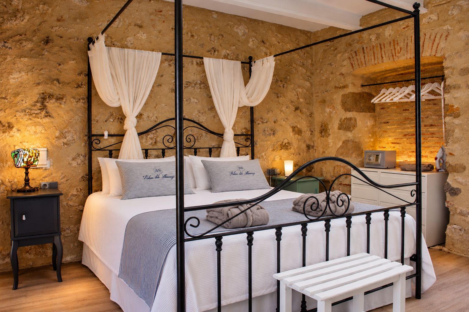 A four-poster bed with a canopy, bedside tables, a bench, and a stone wall background.