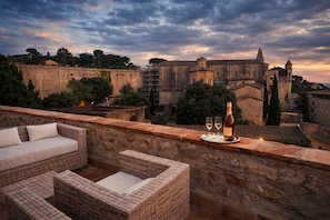A rooftop terrace with wicker furniture, a bottle of wine, and glasses on a tray, overlooking historic buildings and a cloudy sky. - AS Palau dels Alemanys (Girona)