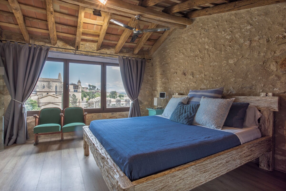 A rustic bedroom with a large bed, wooden beams, stone walls, and a view of a historic building.