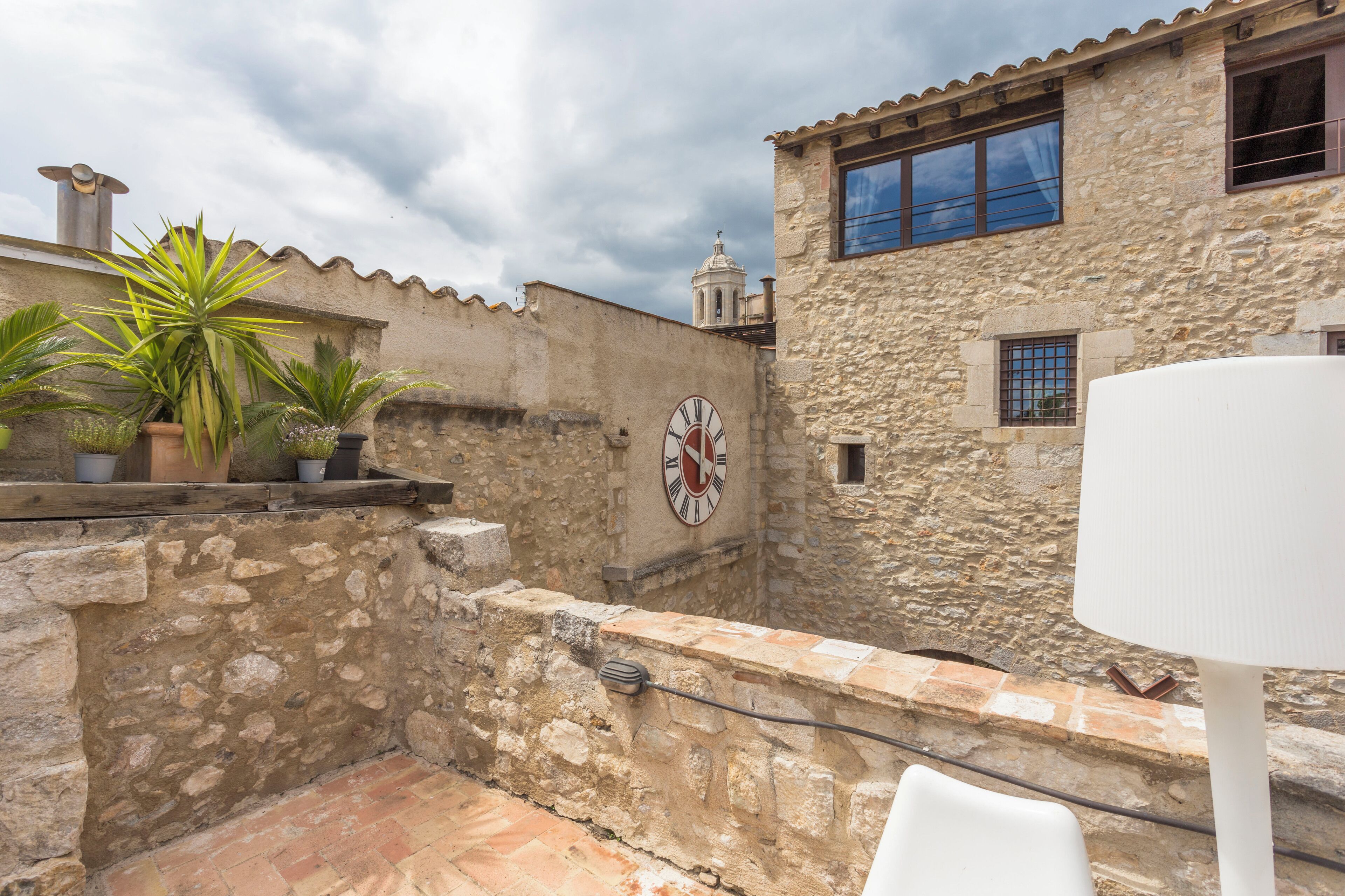 A courtyard with stone walls, a clock, potted plants, and a white table.