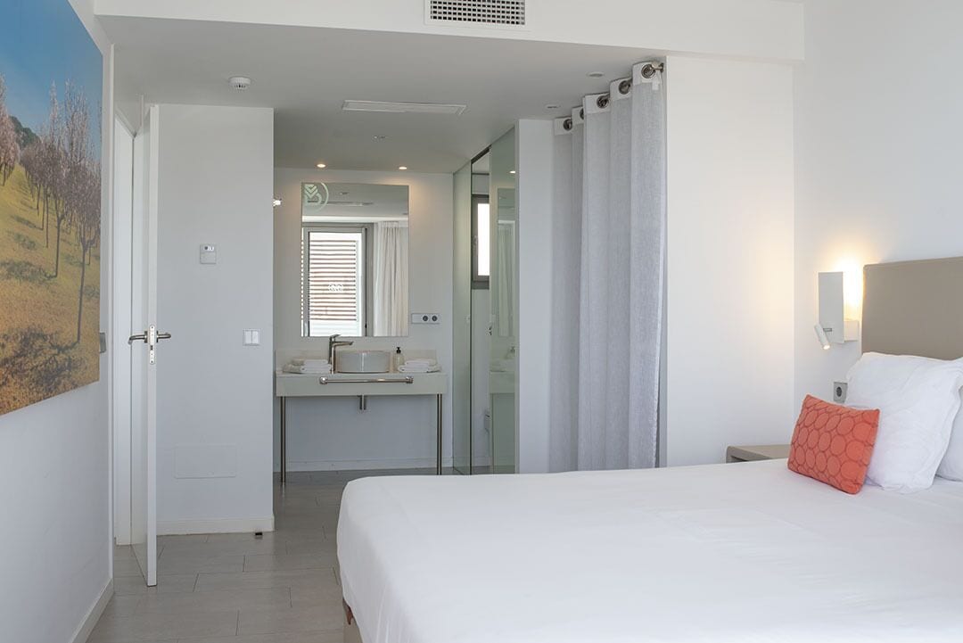 Hotelkamer met een bed, nachtkastje, bedlampje, badkamer met wastafel en spiegel, en een deur naar een andere kamer.