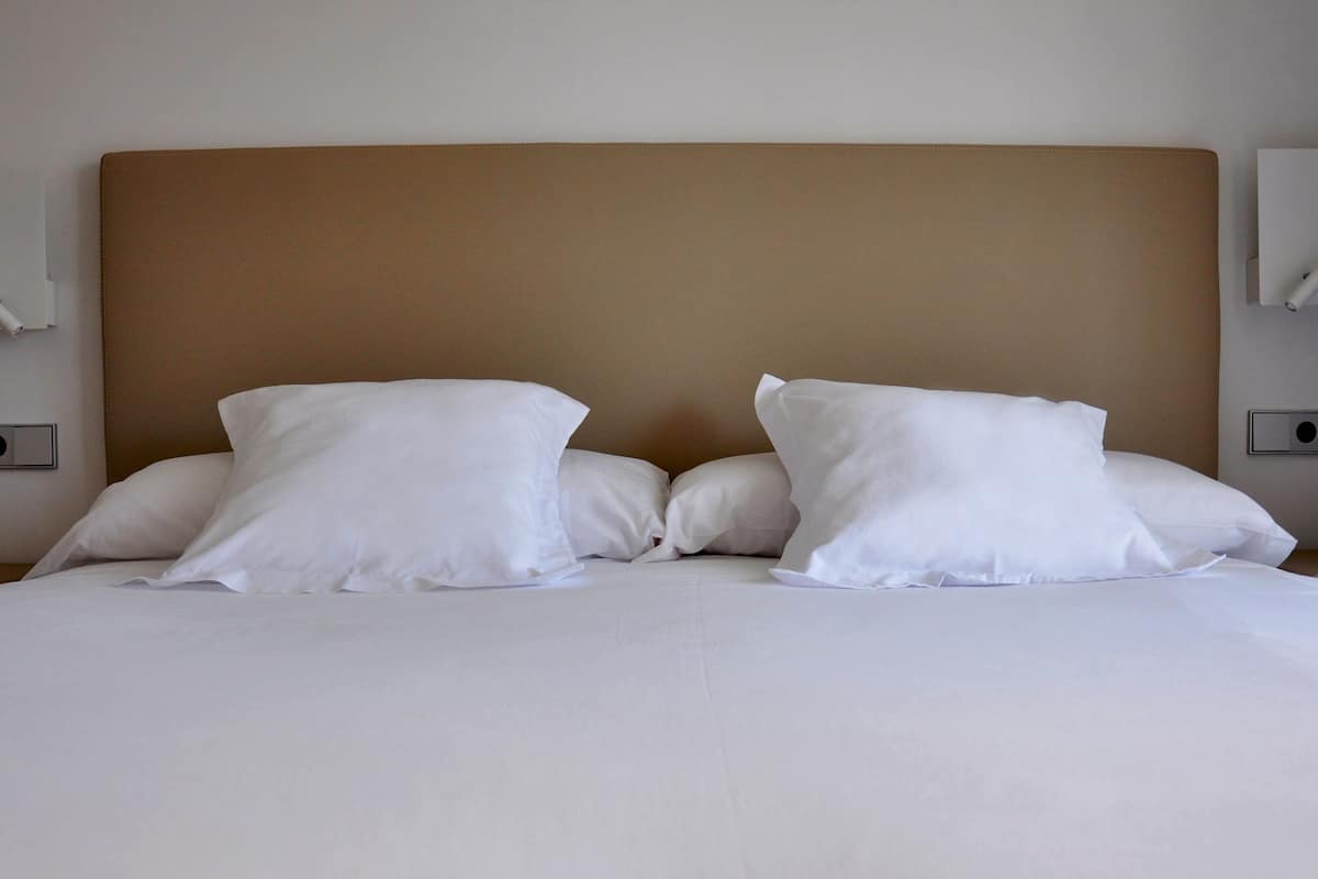 A neatly made bed with white linens and two pillows, a brown headboard, and two wall-mounted light fixtures.