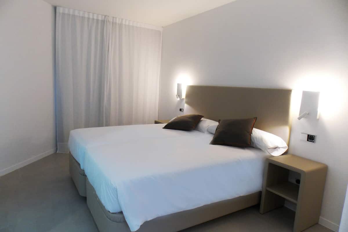 A neatly made bed with white linens and two brown pillows, a bedside table with a lamp, and a window with white curtains.