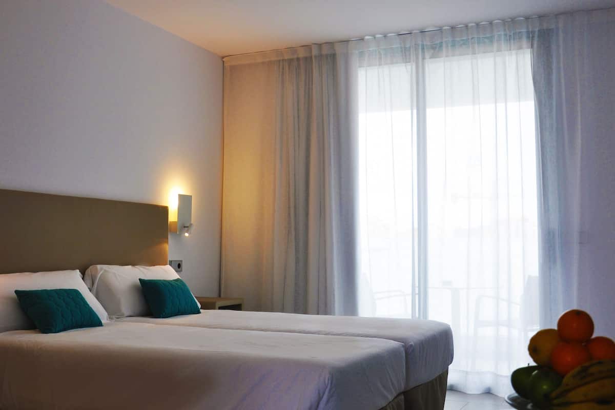A hotel room with two beds, a nightstand with a lamp, a wall-mounted light, and a large window with sheer curtains.