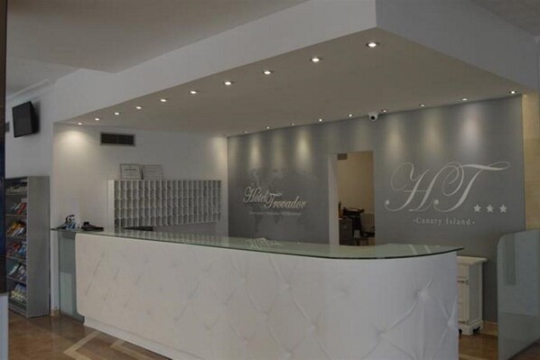 A reception area with a glass counter, a wall-mounted sign reading "Hotel Troador Canary Island," and a ceiling with recessed lighting.