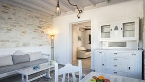 A cozy living room with a stone wall, a white sofa, a dining table with a fruit plate, and a white cabinet with drawers. - Contaratos Beach (Paros)