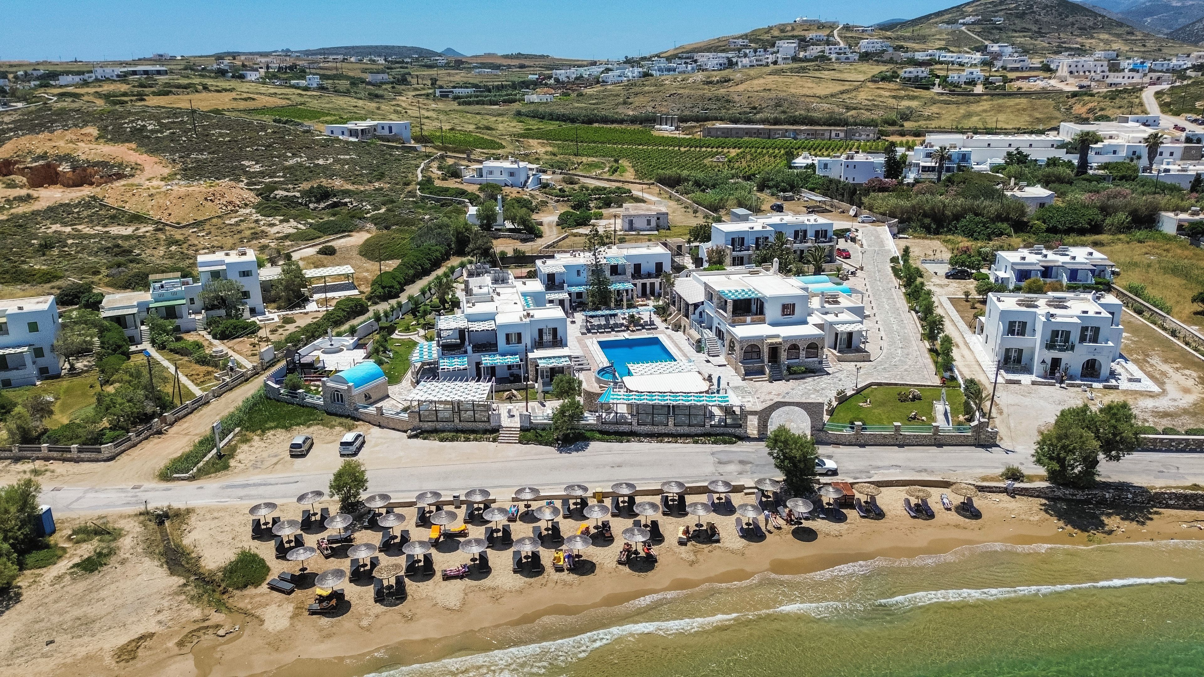 Vista aérea de um resort com vários edifícios, uma piscina central e área de praia com espreguiçadeiras.