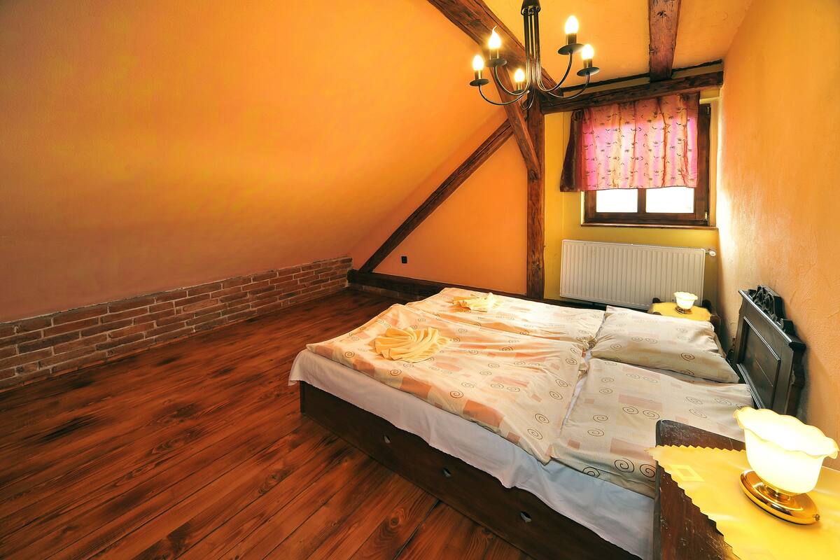 A bedroom with a sloped ceiling, wooden beams, a brick wall, a radiator, a bed with a patterned bedspread, and a nightstand with a lamp.
