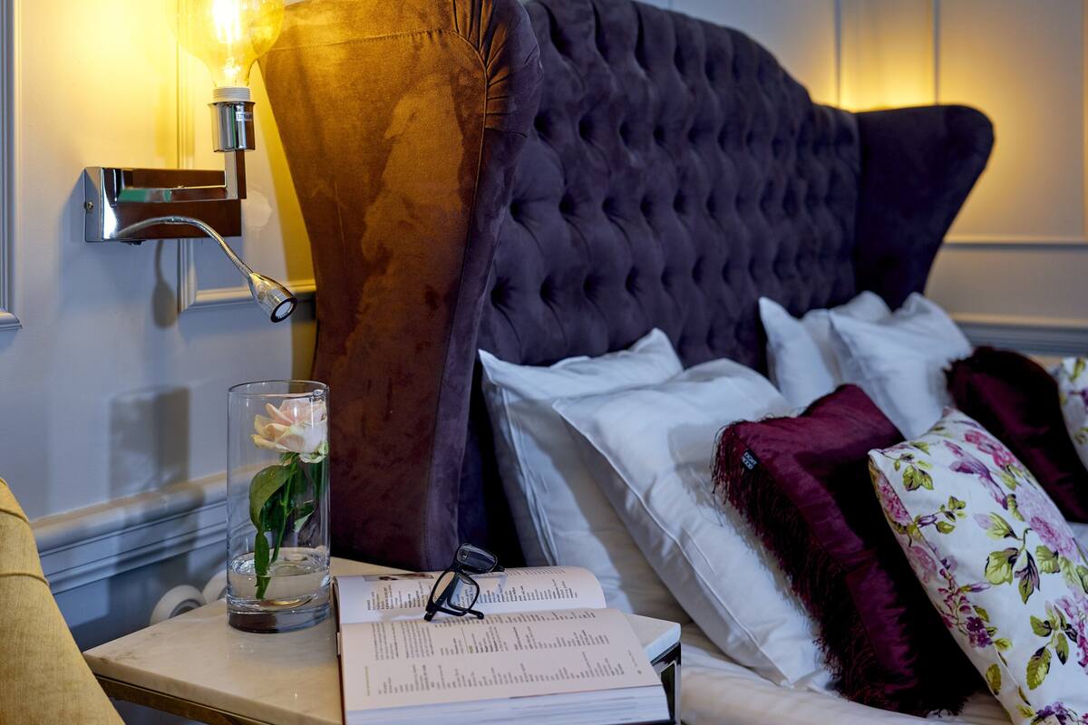 A neatly made bed with white and purple bedding, a glass vase with a single flower, a book, and a pair of glasses on a bedside table.